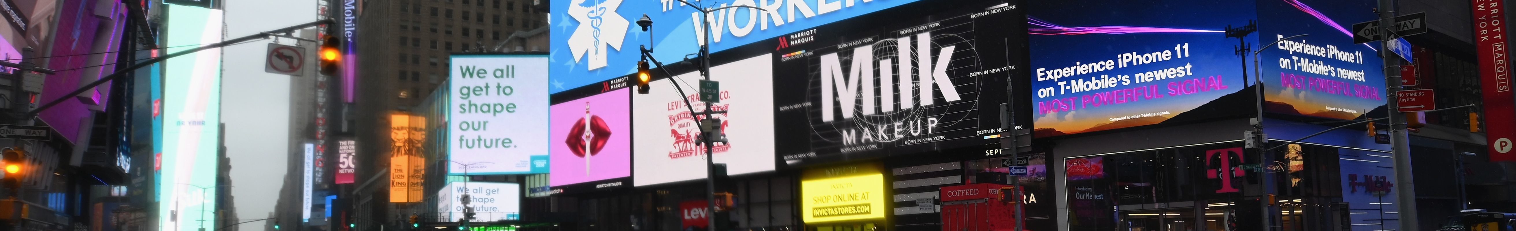 A nearly empty Times Square is seen on March 23, 2020 in New York City. It is raining and the streets are wet. An electronic billboard reads, “Thank You #HealthCare Workers!” Other electronic billboards show ads for various products. The lights from the billboards can be seen reflected in the water on the street.