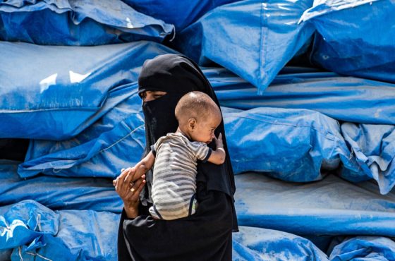 A woman wearing a niqab (full face veil) walks carrying an infant at al-Hol camp for displaced people in al-Hasakeh governorate in northeastern Syria on July 22, 2019, as people collect UN-provided humanitarian aid packages.