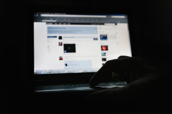 A hand touches a laptop that shows Facebook. Only the screen is lit up; the rest of the photo is dark.