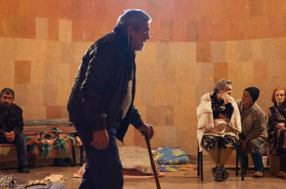 Local elderly residents take shelter in the basement of an undisclosed church on October 12, 2020 in Stepanakert, Nagorno-Karabakh. Boxes and blankets are placed in piles on the floor and there are a few chairs and benches on which people sit. A person walks past the camera using a walking cane.