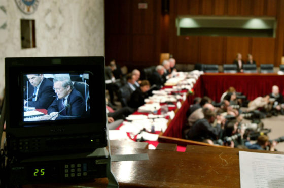 Secretary of Defense Donald Rumsfeld is seen on a television monitor as he testifies before the bipartisan September 11 commission, formally known as the National Commission on Terrorist Attacks Upon The U.S., on Capitol Hill March 23, 2004 in Washington, DC. Photographers can be seen kneeling on the floor with cameras and officials sit at tables around the room.