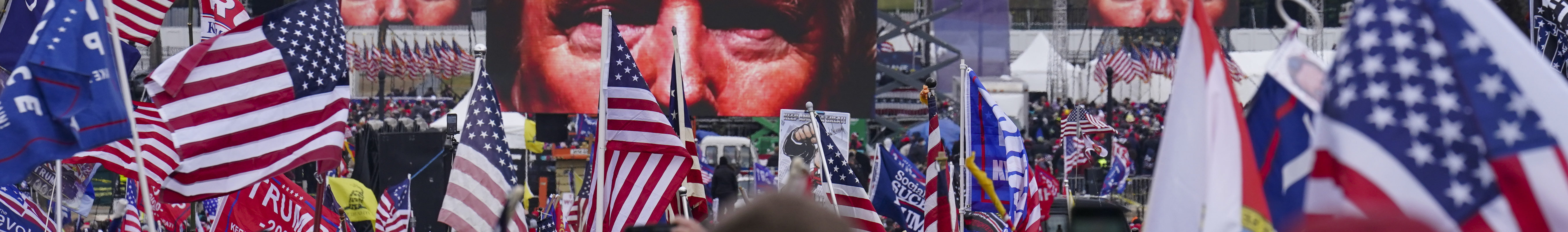 Trump supporters at the January 6th rally at the Ellipse. The Trump propaganda film is playing and three large screens show the top half of Trump’s face under an ominous red light. People in the crowd wave American flags and Trump 2020 flags.