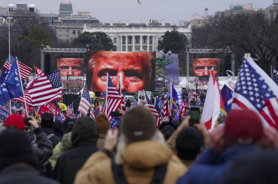 Trump supporters at the January 6th rally at the Ellipse. The Trump propaganda film is playing and three large screens show the top half of Trump’s face under an ominous red light. People in the crowd wave American flags and Trump 2020 flags.