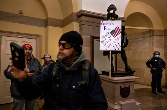 Trump supporters infiltrate the US Capitol on January 6, 2021, in Washington, DC. They do not wear face masks, and one man appears to take a selfie or video on a phone. The insurgents placed a sign against a statue. The sign reads, “America First Never Give up Never Surrender.” A police officer, who wears a surgical mask, appears to stand nearby watching them but not stopping them.