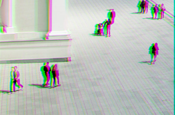 People walk in a courtyard near a tall building. The colors of the image are separated and blurred in a psychedelic camera effect to give the appearance of seeing double.