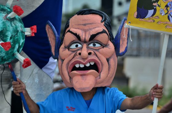 A medical doctor wearing a mask representing Honduran President Juan Orlando Hernandez takes part in a protest in Tegucigalpa on September 11, 2020, amid the new coronavirus pandemic. The doctor carries a 3-D coronavirus model in one hand and a sign in another hand.