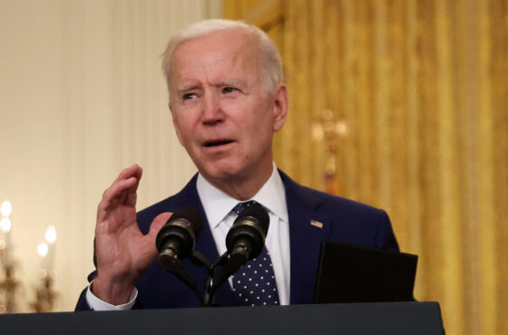 U.S. President Joe Biden stands at a podium and announces new economic sanctions against the Russia government from the East Room of the White House on April 15, 2021 in Washington, DC. A chandelier-type lamp and multiple flags stand behind him.