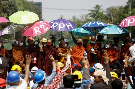 Monks take part in a demonstration against the military coup in Yangon on March 11, 2021. They carry umbrellas reading, “R2P” standing for the Responsibility to Protect and “CRPH” standing for the Committee Representing Pyidaungsu Hluttaw. The demonstrators wear face masks. Other demonstrators wear hard hats and raise three fingers in the air.