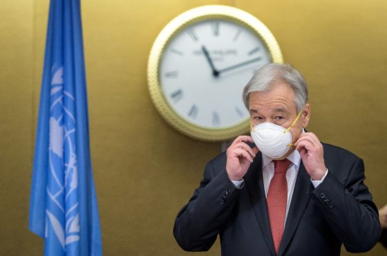 UN Secretary General Antonio Guterres removes his protective facemask prior to attending a press conference, following the 5+1 meeting on Cyprus, in Geneva, on April 29, 2021. Behind him is a clock and the UN flag.