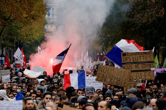 Protesters wave French flags and signs written in French as they march near the Gare du Nord, in Paris to protest against Islamophobia, on November 10, 2019. A cloud of smoke rises behind them.