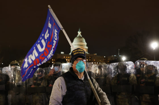 An insurrectionist with a MAGA hat and Trump flag stands in front of the national guard outside the Capitol building the evening of January 6th.